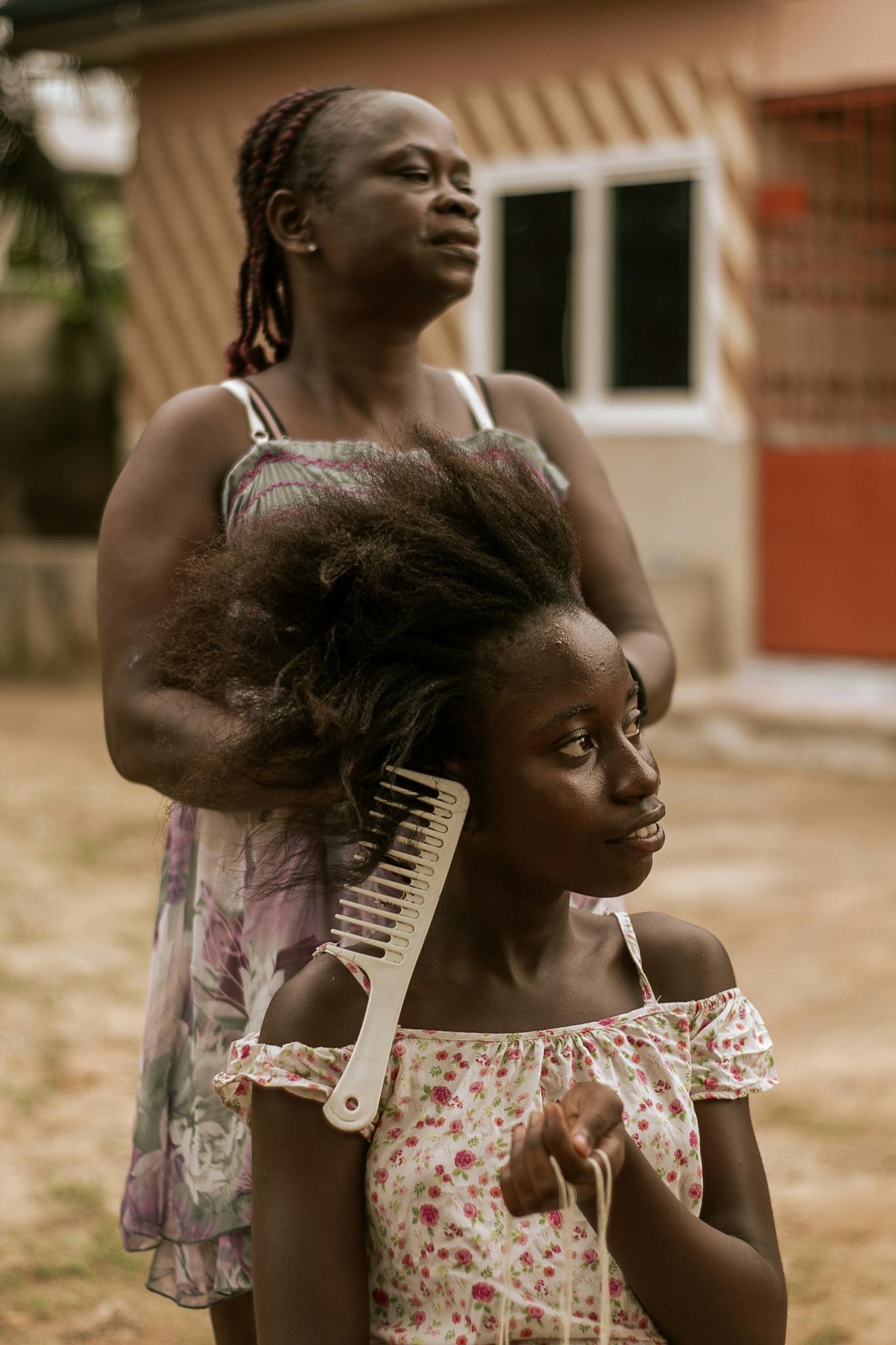 A mother combs her daughter's hair outdoors, capturing a tender bonding moment.