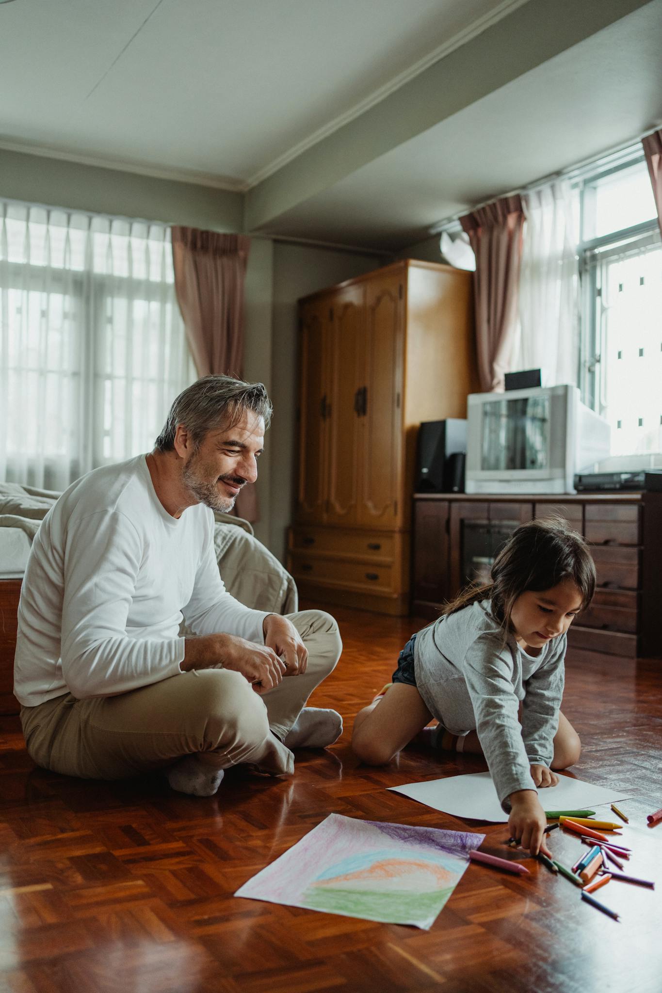 Father and daughter sharing joyful moments while coloring together on a wooden floor.